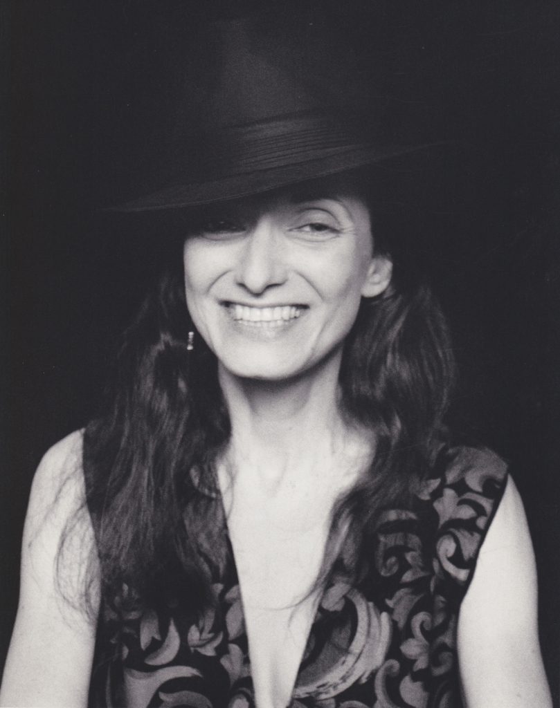 Black and white image of photographer Flo Fox. In this image, Flo wears a black fedora has long dark hair and is wearing a sleeveless printed top. The portrait is bust length.