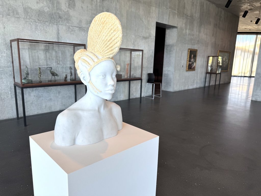 A white marble bust of a person with an elaborate, golden braided hairstyle stands on a white pedestal within a modern, concrete-walled museum gallery space.