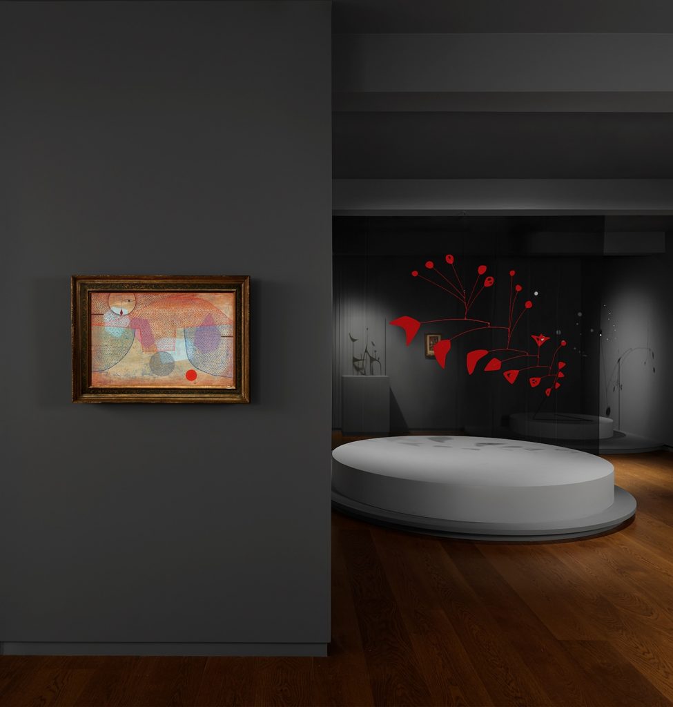 an installation view of a dimly lit gallery with a colorful abstract painting hanging in the foreground and a red sculptural mobile in the background
