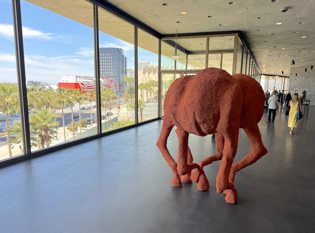 A large, textured terracotta-colored sculpture of abstract animal legs stands in a bright museum gallery. Floor-to-ceiling windows reveal a sunlit city street with palm trees.