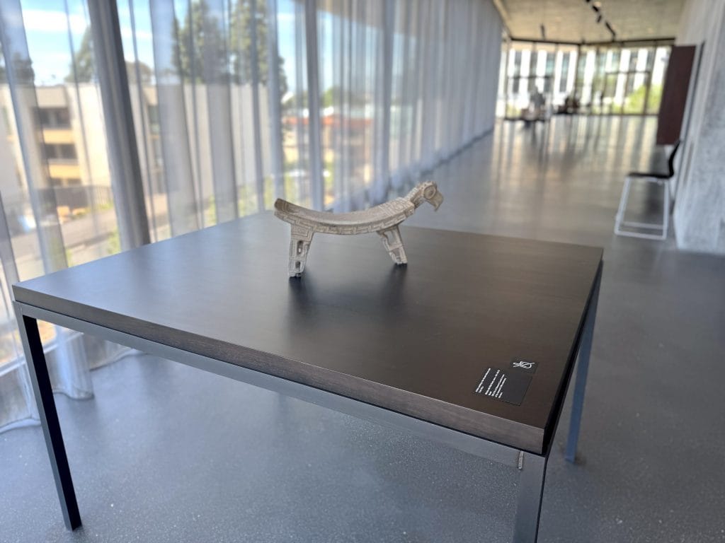 An ancient stone metate with a carved bird head sits on a dark display table in a long, sunlit gallery lined with sheer window curtains.