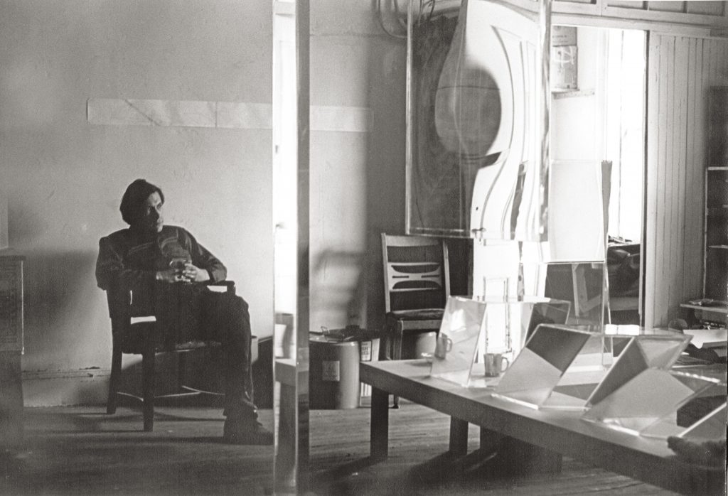 Black-and-white photograph of an artist seated in a studio, surrounded by geometric glass sculptures and reflective panels.