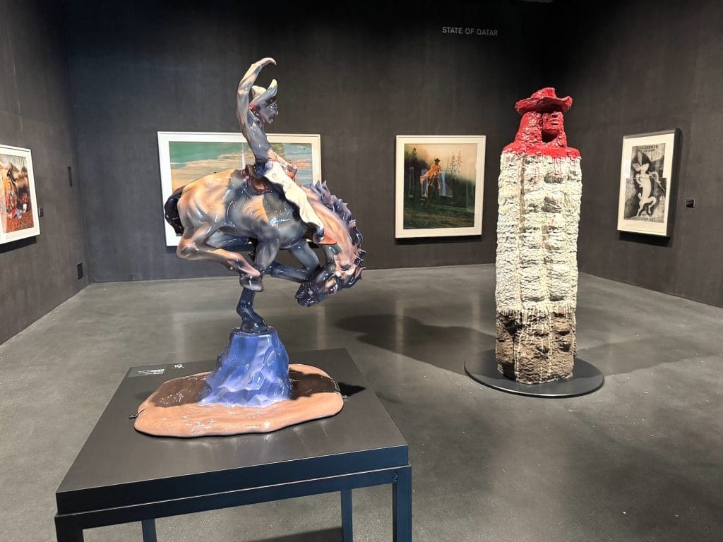 A glossy sculpture of a bucking bronco and a tall, textured figure with a red hat stand in a dark gallery featuring Western-themed artworks.v