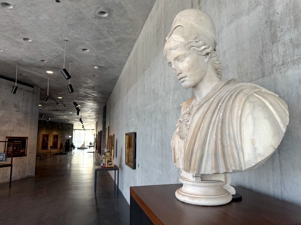 A classical marble bust of Athena wearing a helmet is shown in profile against a concrete wall