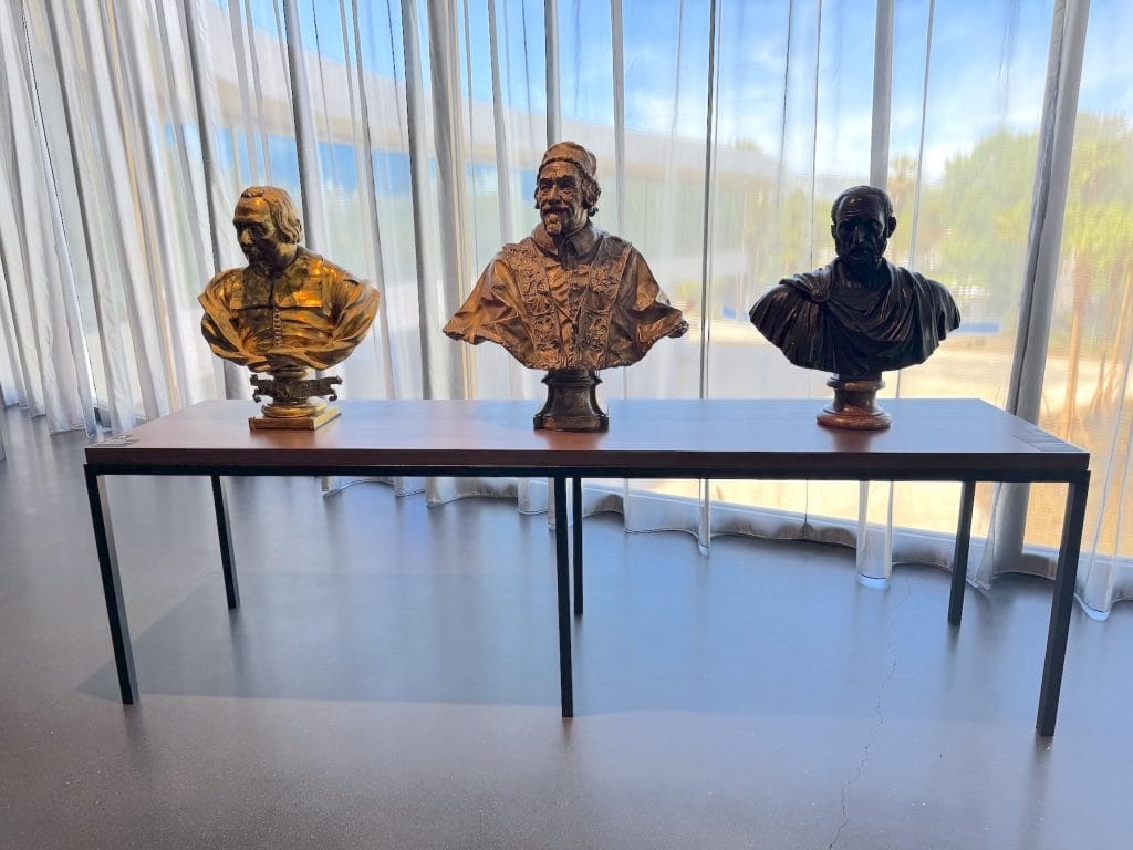 Three bronze and gold-toned portrait busts of men are displayed on a wooden table in front of floor-to-ceiling windows with sheer, light-filtering curtains.