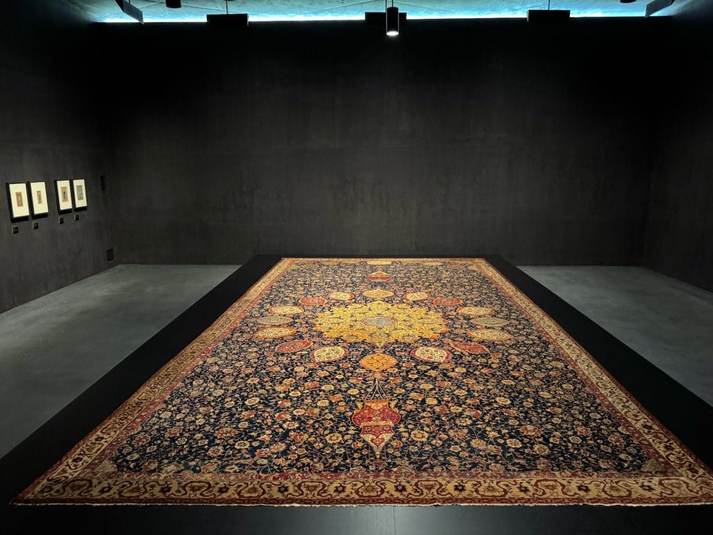 A large, intricately patterned Persian carpet with a central golden medallion and floral motifs is displayed on a low black platform in a dark gallery.