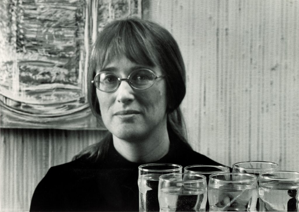 A black and white photo of a young white woman in a black shirt seen from the shoulders up, with water glasses in the foreground and a large still life painting of glasses hanging on the wall behind her. She has her hair pulled back into a loose ponytail with bangs and is wearing thin rimmed glasses. 