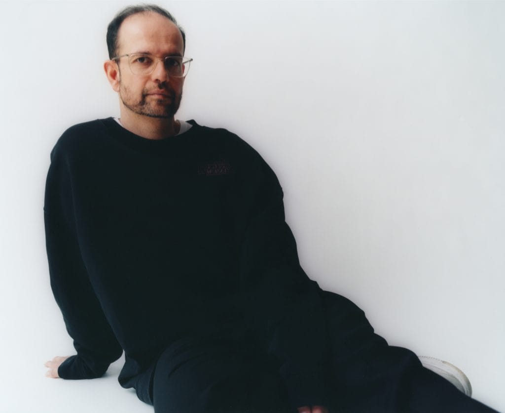 Portrait of artist Tino Sehgal seated against a white wall, wearing a black sweatshirt and glasses, looking directly at the camera.
