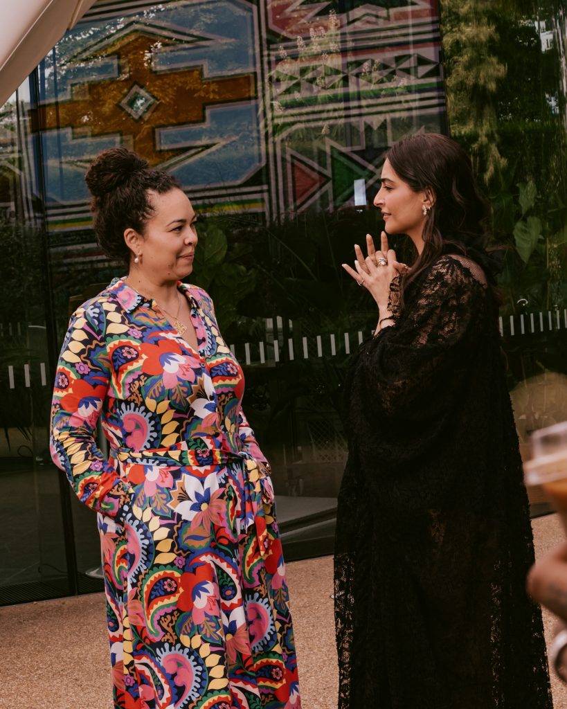 Two women engaged in conversation at a cultural event, one wearing a vibrant floral-patterned dress and the other in a black lace outfit, standing against a backdrop of colorful geometric wall art.