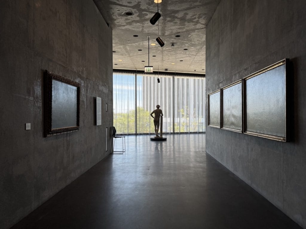 A concrete museum hallway features framed paintings on both walls, leading toward a silhouette of a classical male statue standing before a bright, curtained window.