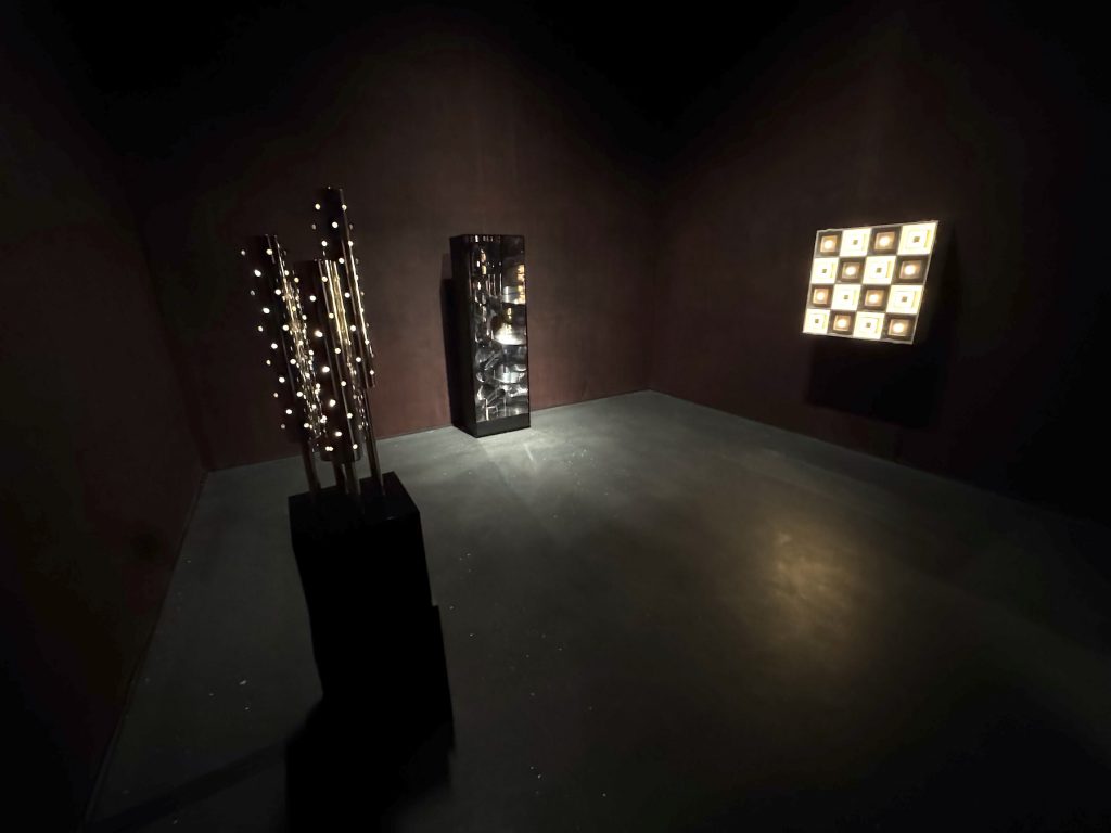 Three kinetic and light-based sculptures illuminate a dark gallery. The works feature metallic cylinders with small lights, a reflective pillar, and a glowing geometric grid.