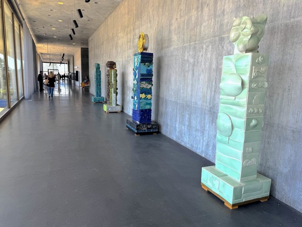 A row of tall, colorful ceramic totems by Dora de Larios lines a concrete gallery wall, leading toward a sunlit window at the hallway's end.