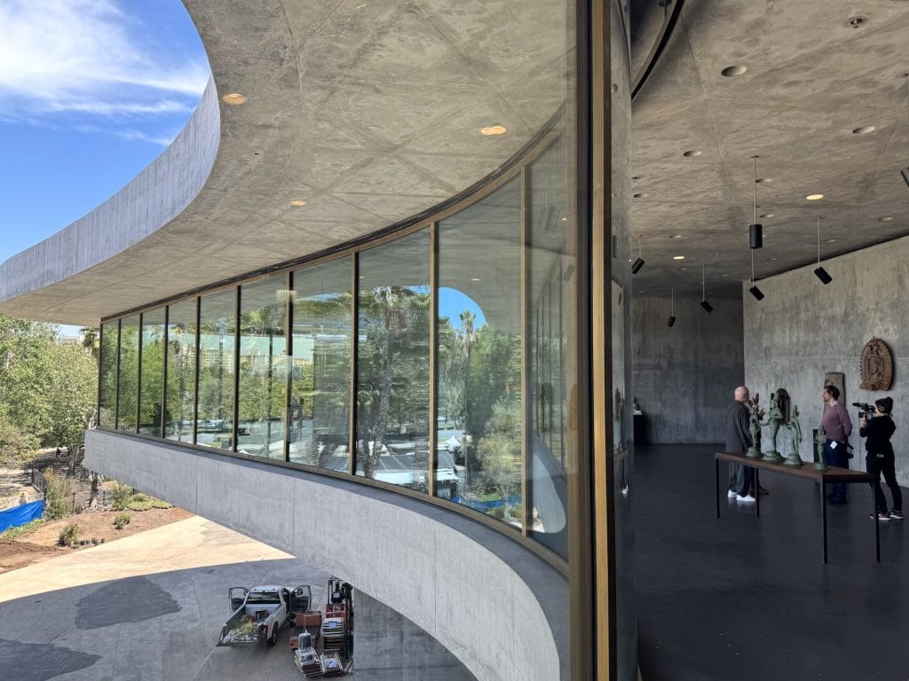 A curved concrete gallery features floor-to-ceiling windows reflecting palm trees. Inside, visitors observe small bronze sculptures on a table against a raw concrete interior wall.