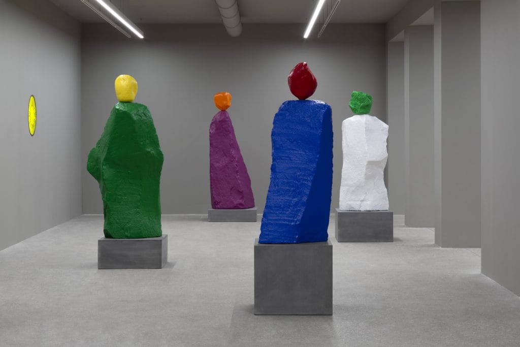 A gray-walled room filled with colorful rocks sitting on grey plinths