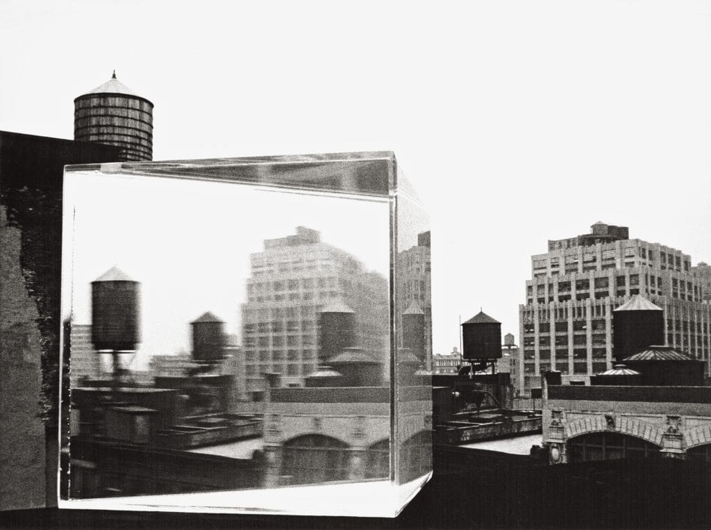 A glass cube on a rooftop reflecting blurred city buildings and water towers against an urban skyline.