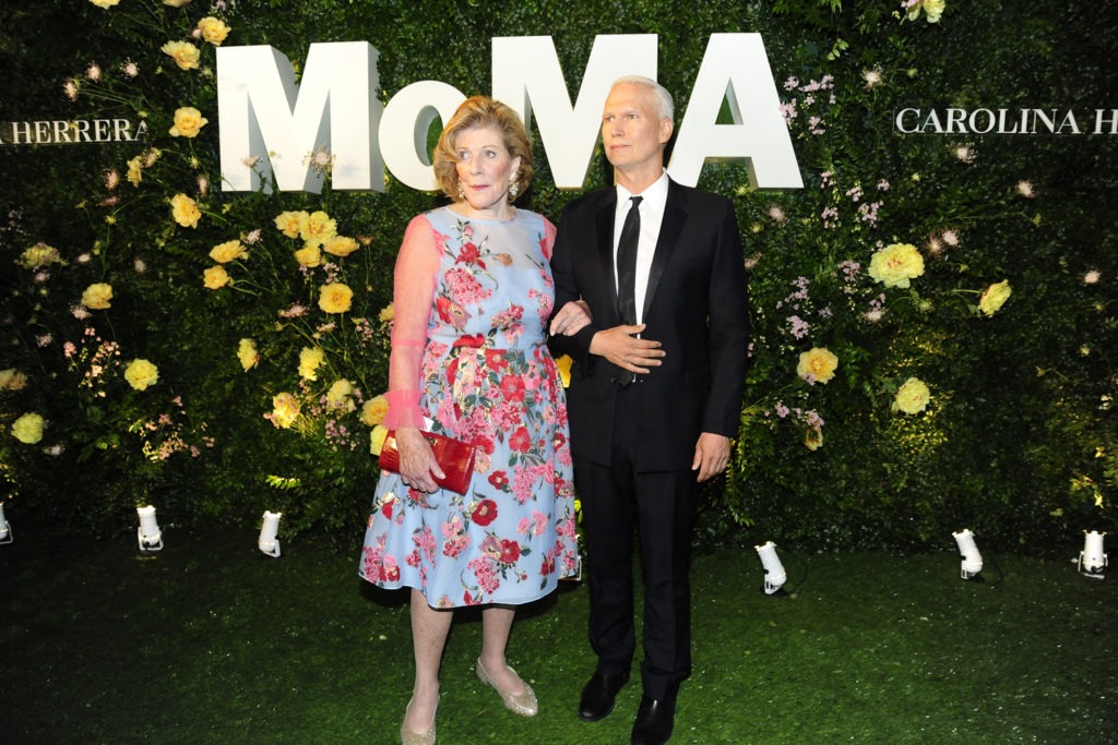 A photo of an older white woman in a light blue floral dress with sheer pink sleeves, holding a red clutch, standing arm in arm with an older white man in a black suit and tie. They pose in front of a green wall decorated with yellow flowers and a large “MoMA” sign.