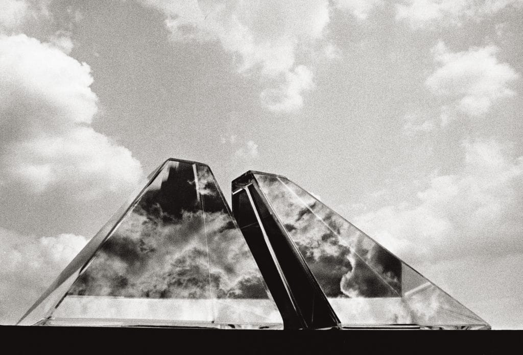 Two pyramid-like glass structures reflecting clouds in a bright sky, creating mirrored geometric forms.