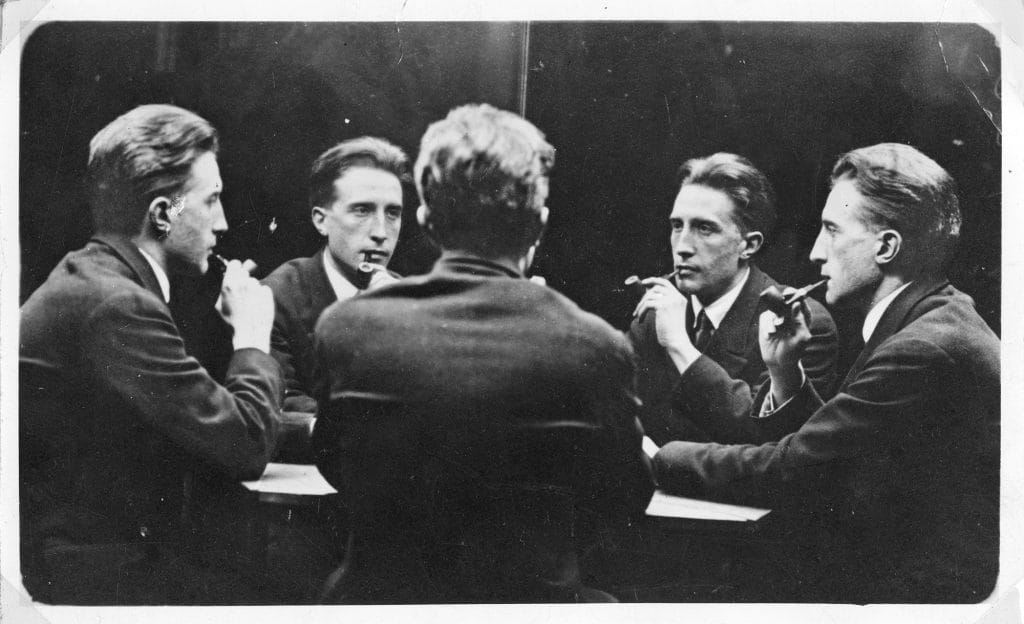 Black-and-white photograph of a man seated at a table, reflected multiple times in mirrors to create five views of his face.