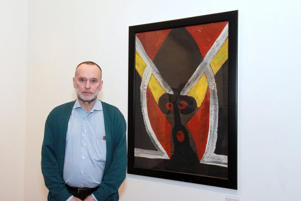 Man standing beside an abstract painting with bold red, yellow, black, and white shapes in a gallery setting, highlighting contemporary art exhibition photography.