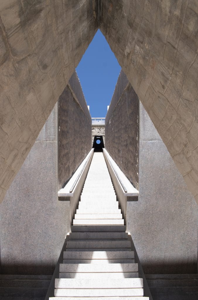 A narrow stone corridor with stairs leading toward a circular opening framing the sky.