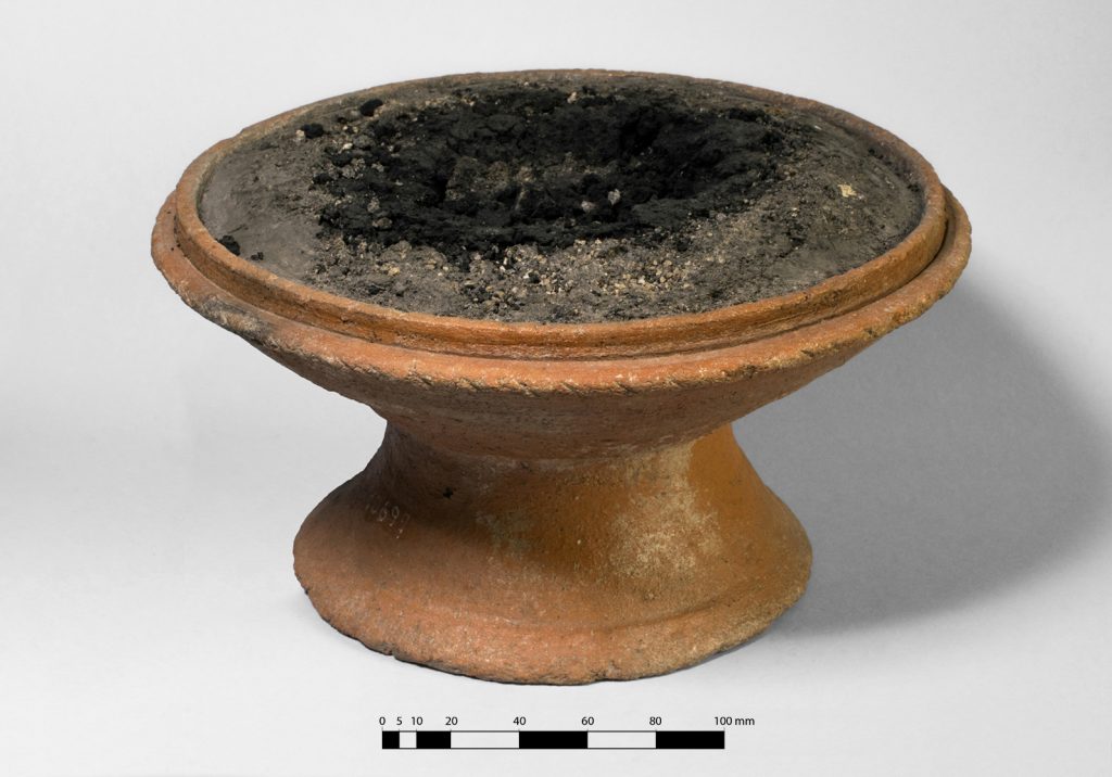 A photograph of a small clay vessel full of burnt gray material in it atop an all-white background