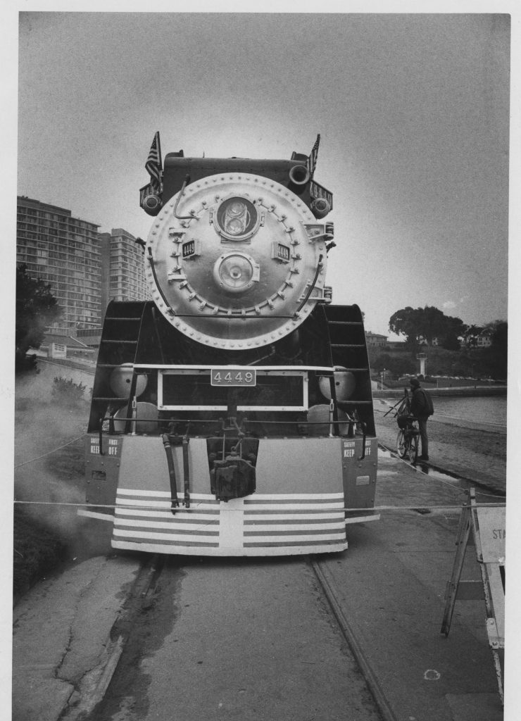 A black and white photo of the front of a large steam locomotive numbered 4449 stopped on railroad tracks, with steam drifting around the base. Tall apartment buildings and trees appear in the background, and a man stands beside a bicycle near the tracks.