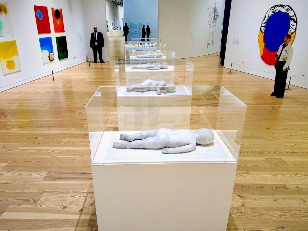 Five life-size wax sculptures of nude toddlers by Andrea Fraser are displayed on white pedestals inside transparent Plexiglas cases against a large gallery window.