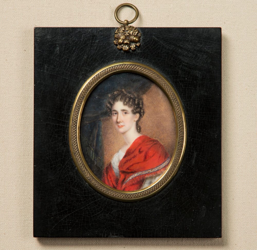 a painting of a young white woman with curly dark hair, wearing a red shawl over a white dress, shown in a small oval miniature set within an ornate black and gold hanging frame