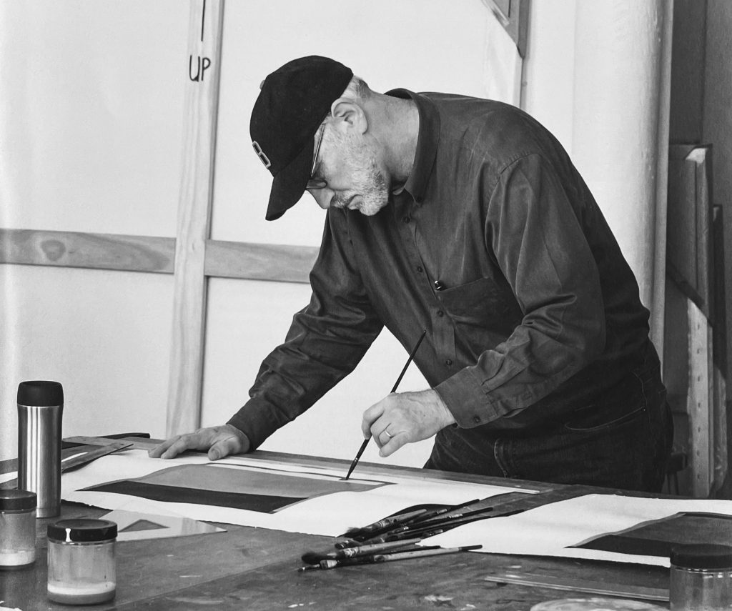 Black-and-white photo of an artist in a studio carefully painting geometric shapes on paper, surrounded by brushes and jars that highlight the precision and craft of abstract art making.