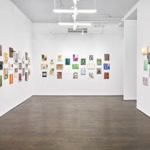 A photo of a white cube gallery space with rows of small, colorful rectangular paintings hung on the wall in a neat grid.