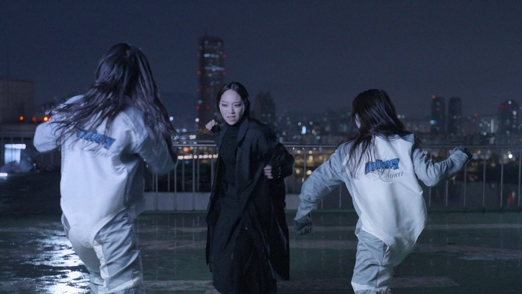 A video still from an Ayoung Kim film featuring two women in white suits lunching towars another woman in all black, in a vast concrete space amidst a nighttime city scene