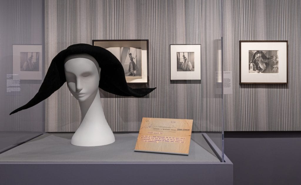 Installation view featuring a sculptural black hat displayed on a white mannequin head in a glass case, with framed black-and-white fashion photographs by Lillian Bassman hung on a striped wall behind.