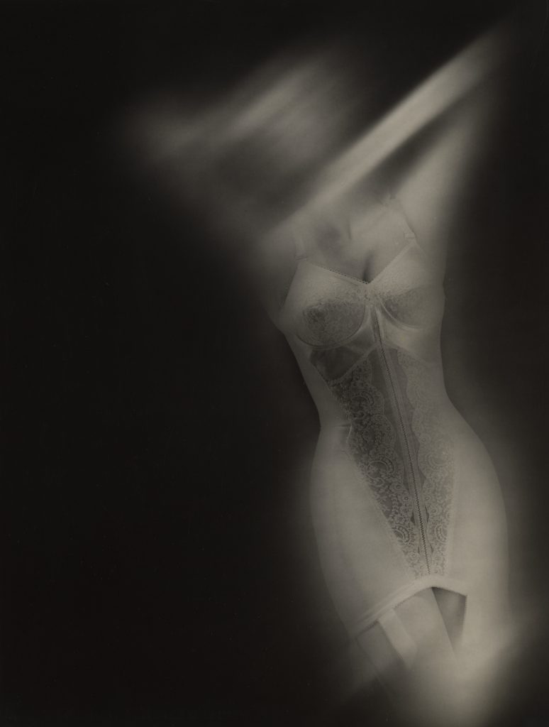 Soft-focus photograph of a female figure in a lace bodysuit, partially veiled by blurred light streaks that obscure her face and upper body, emphasizing the contours of the garment.
