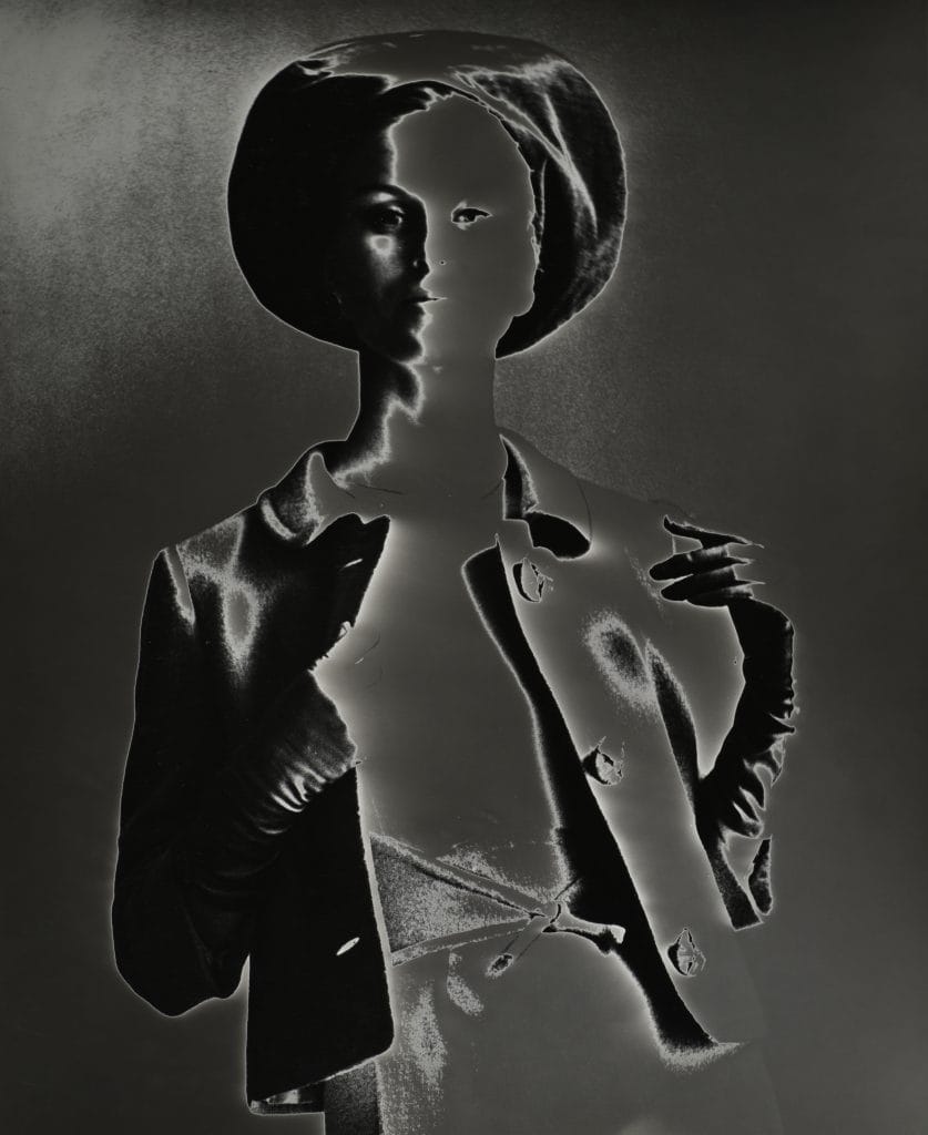 High-contrast, solarized photograph of a female model wearing a hat and jacket, her face partially obscured by negative-like inversion, creating a stark, glowing silhouette against a dark background.