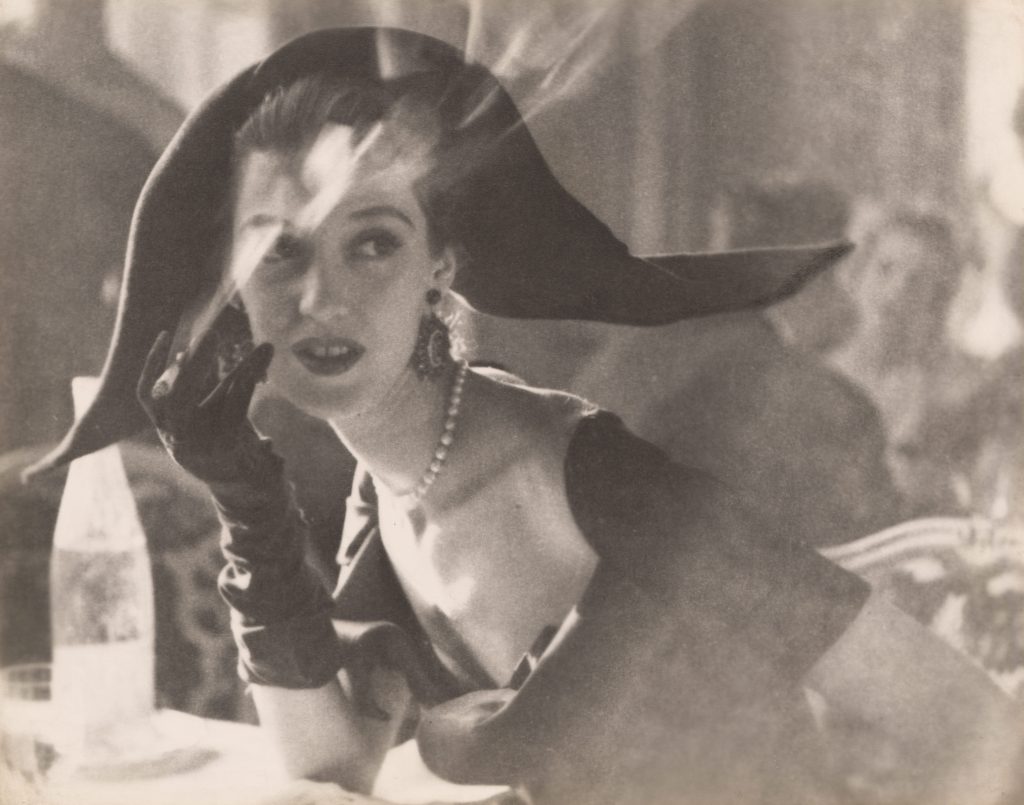 Soft-focus black-and-white photograph of an elegantly dressed woman wearing a wide-brimmed hat, gloves, and a pearl necklace, seated at a table and touching her face, with blurred figures and light reflections in the background.