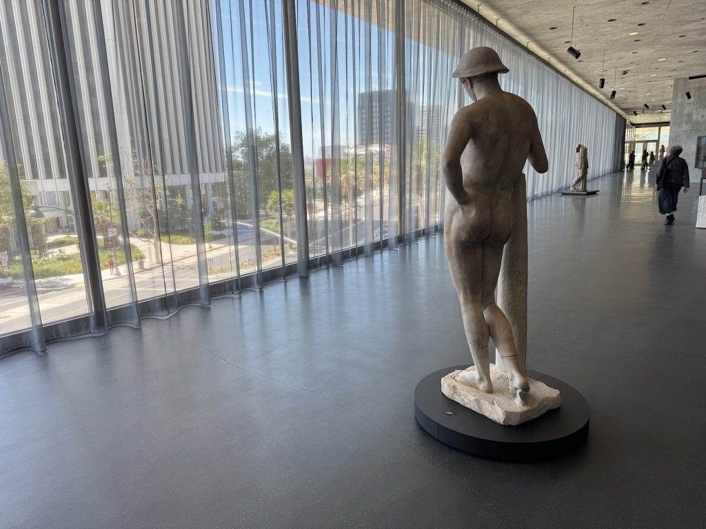 A silhouette of a classical male sculpture stands before floor-to-ceiling windows with sheer curtains, overlooking a sunlit city street and lush greenery in a museum.