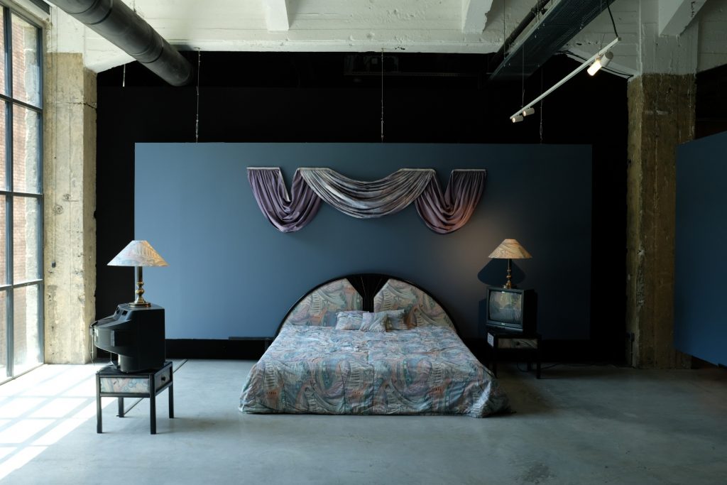 an installation made to look like a typical bedroom from the 1990s with a bed and some other pieces of domestic furniture including side tables and lamps 