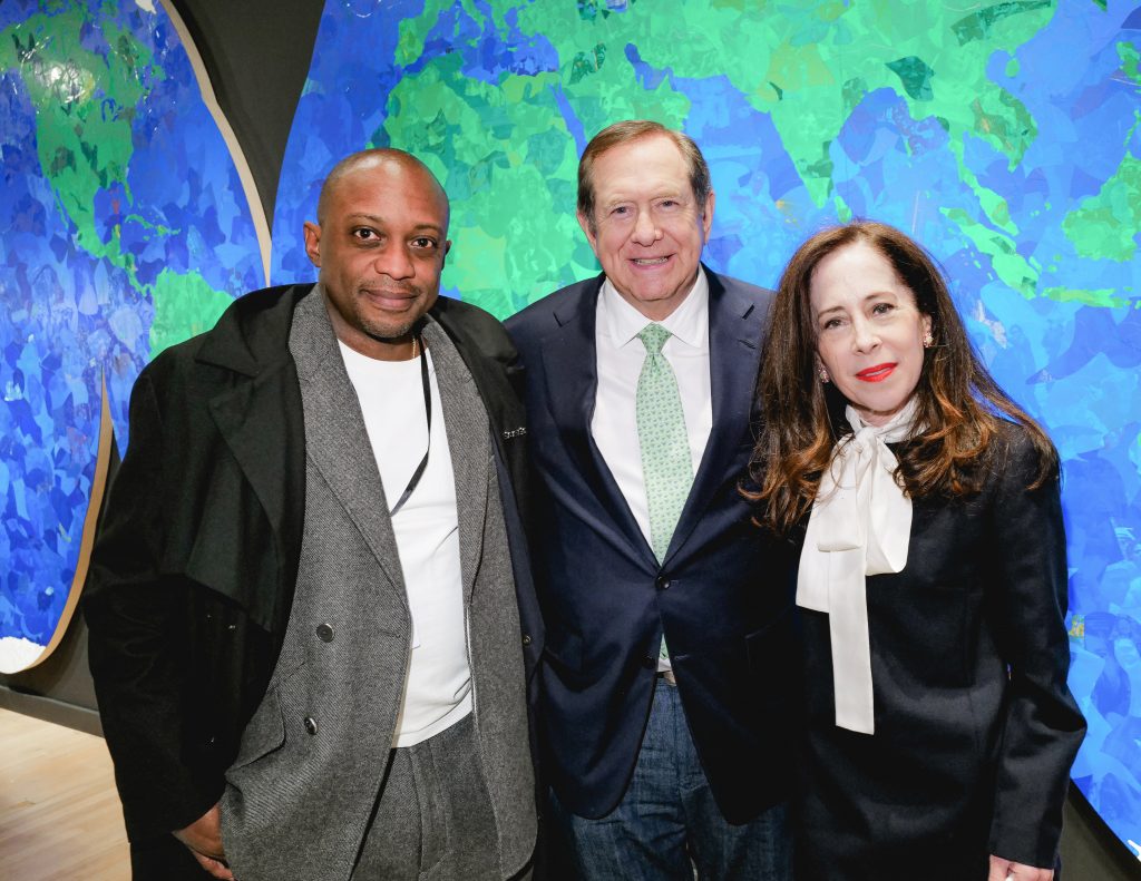 Hank Willis Thomas, Jordan Schnitzer, and curator Sharon Coplan pose together in front of a large, colorful, map-like artwork in layered blue and green tones.