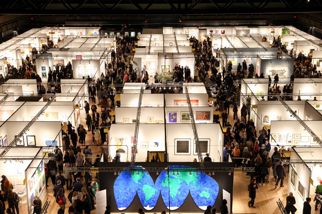 A wide view of the IFPDA Print Fair filled with booths and crowds, with Hank Willis Thomas’s large blue world map artwork prominently displayed in the foreground.