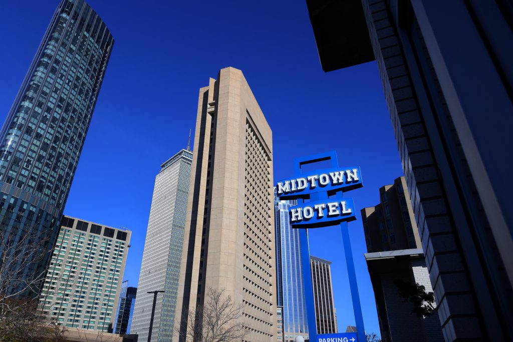 A photo of tall downtown skyscrapers viewed from below under a deep blue sky, with a prominent blue sign reading “Midtown Hotel” in the foreground and modern high-rise buildings surrounding it.