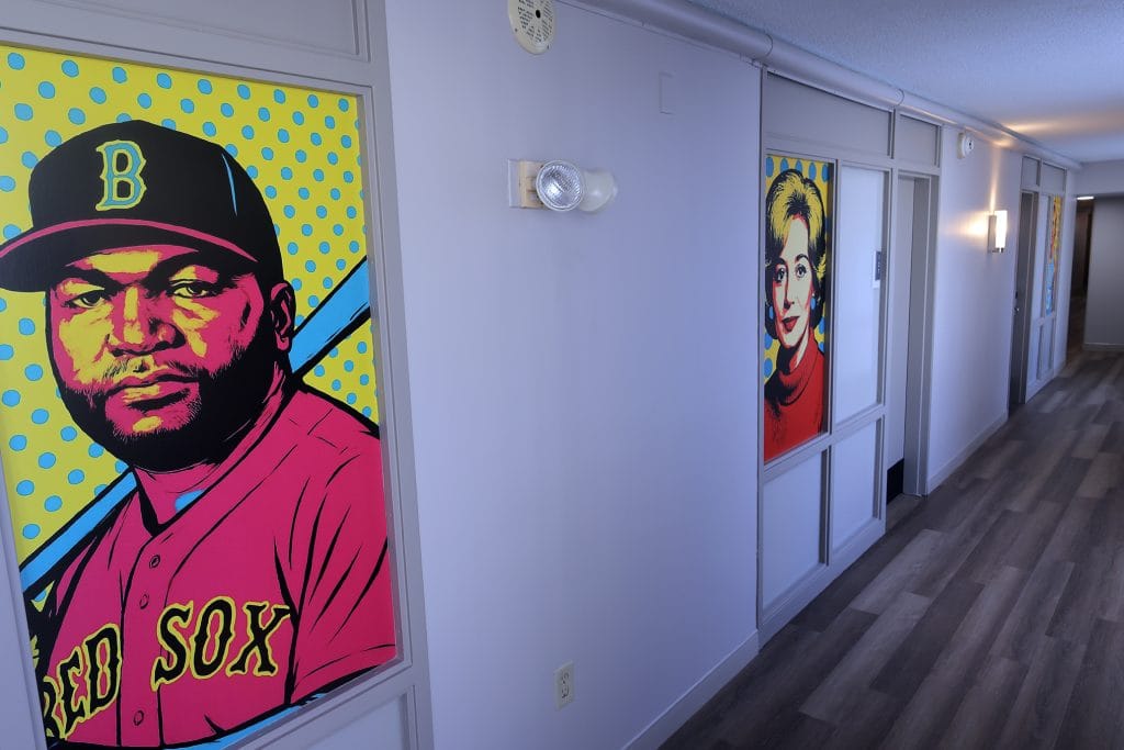 A photo of a hotel hallway featuring large pop-art style portrait murals of adults of different races and genders mounted on the walls, including a Black adult man in a baseball uniform and a white adult woman with short blonde hair, set against bright, dotted backgrounds along a long corridor.