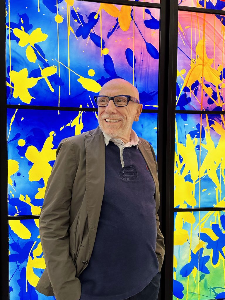 Smiling man wearing glasses and a casual jacket standing in front of a vibrant blue, yellow, and pink abstract artwork with bold splashes and floral silhouettes.