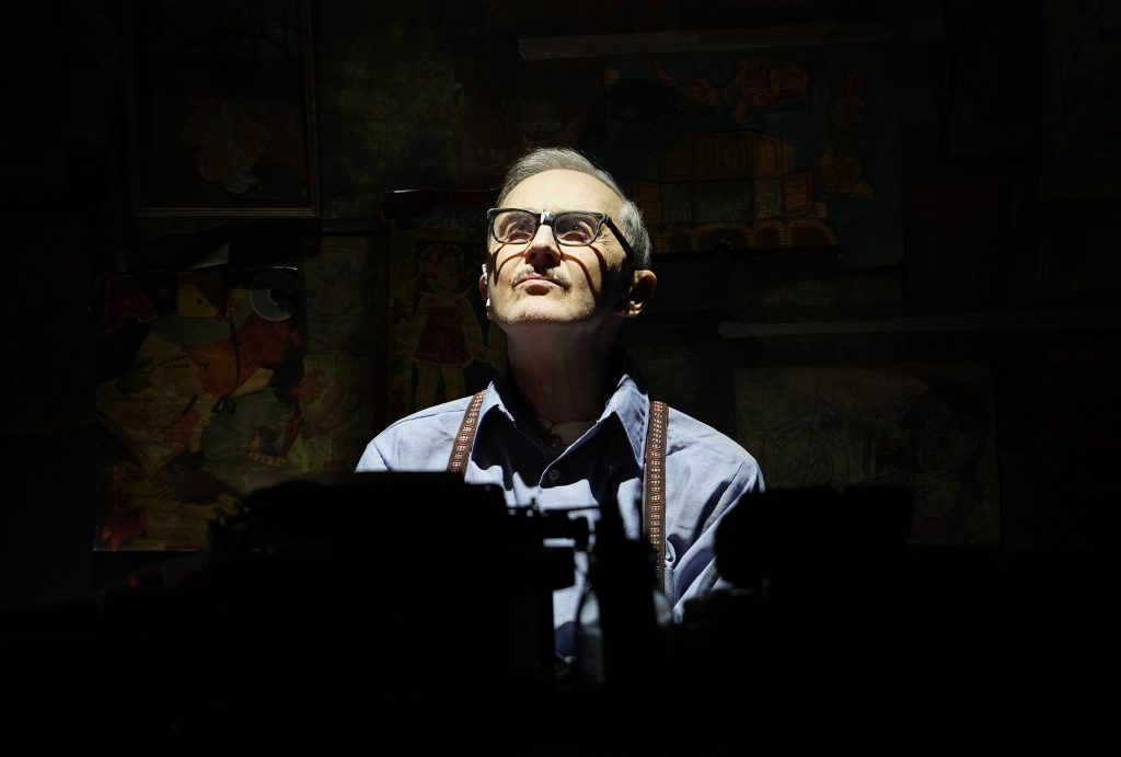 Man in suspenders and glasses gazes upward, face illuminated against dark, shadowy background.