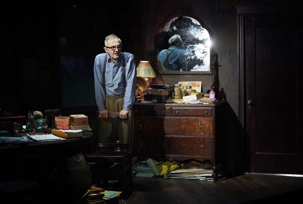 Man leans on chair beside cluttered dresser and mirror reflecting bright burst of light.