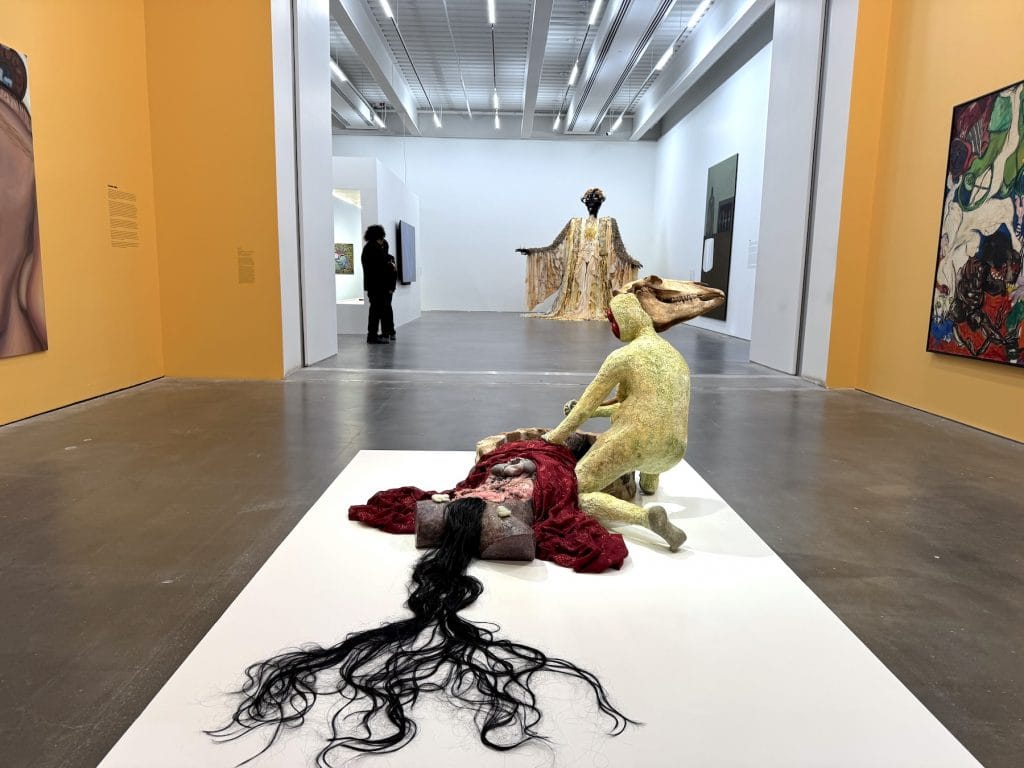 A pale, sculptural figure with an animal skull head kneels over a torso draped in red fabric and long black hair in a brightly lit gallery.
