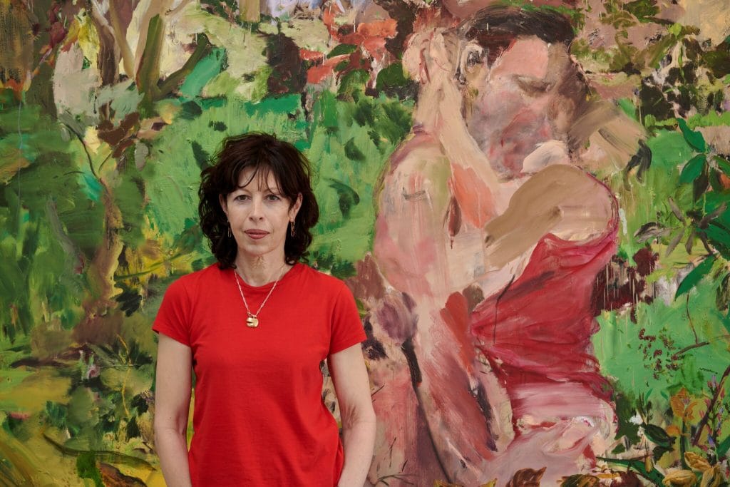 a woman in a red t-shirt stands in front of a painting of a couple locked in an embrace