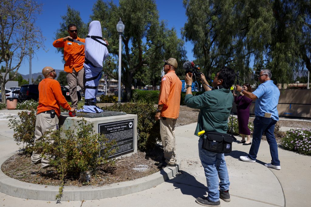 Workers cover Cesar Chavez statue with cloth as onlookers and media document removal in park