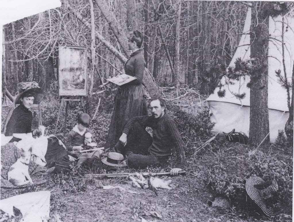 A black-and-white photograph of a group of people seated outdoors in a field, painting on easels with large rock formations in the background.