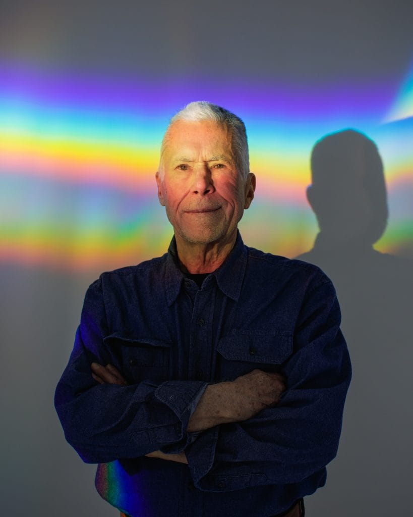 Portrait of an older man with short gray hair standing with arms crossed, illuminated by a band of rainbow light projected across a neutral background.