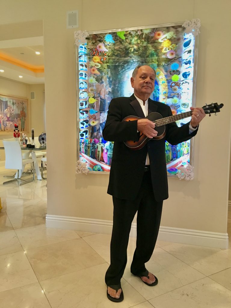 Cheech Marin playing ukulele in front of a colorful contemporary artwork at home, dressed in a black suit and flip-flops, reflecting his passion for Chicano art and eclectic collecting style.